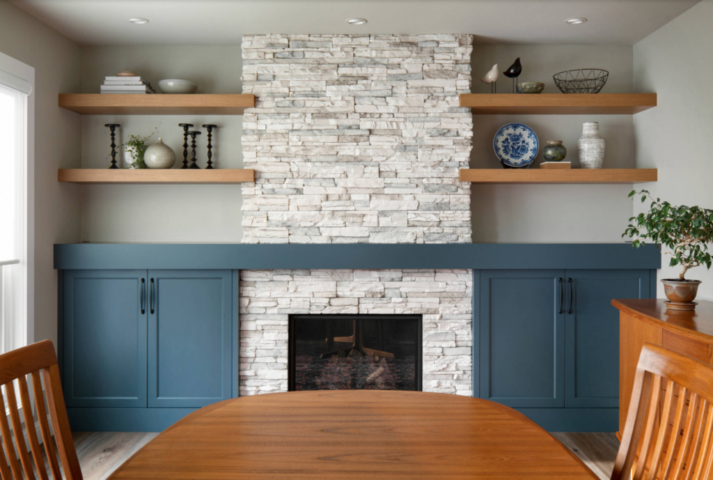 Stone fireplace in a dining room that has floating shelves nearby and blue painted cabinets.